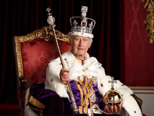 Foto oficial do Rei Charles III, com uma coroa na cabeça, segurando um item, sentado em um trono vermelho e dourado, sorrindo para a câmera e ocm um fundo neutro ao fundo.