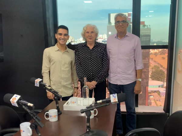 Matheus Andrade, jornalista; ao lado de Dulce Rita, vereadora de São José dos Campos; e Hélcio Costa, também jornalista; após entrevista; na foto os três sorriem para a câmera em frente a uma janela e uma parede preta, com uma mesa de madeira à frente.