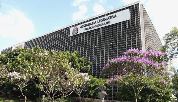 Fachada da Alesp, com uma placa branca colada no prédio, com árvores na frente e um céu azul; foto ilustra matéria sobre o novo salário mínimo paulista.