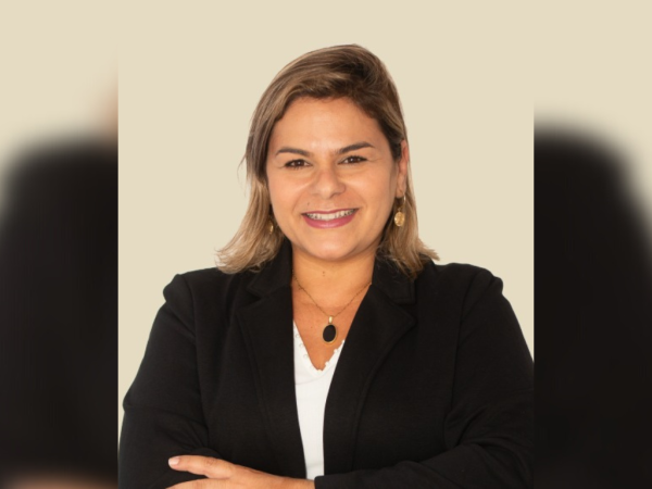 Flávia Pascoal, prefeita de Ubatuba, com um terno preto, camisa branca, sorrindo, com os braços cruzados e em frente a uma parede branca.
