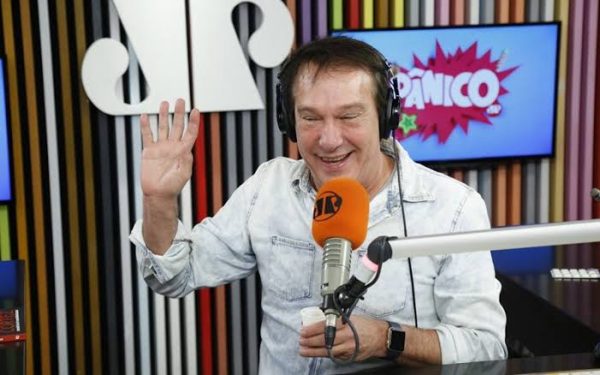Emílio Surita, apresentador do programa Pânico, com uma blusa preta, usando fones de ouvido, olhando para o lado esquerdo, com um microfone em sua frente; foto ilustra matéria sobre seu estado de saúde.