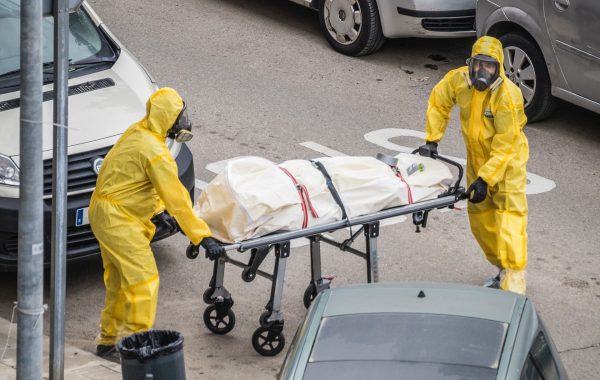 profissionais da saúde com macacões amarelos e máscaras de proteção carregando corpo envolto em tecido branco em maca na rua durante a pandemia da covid-19