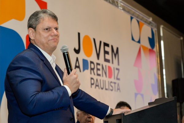 Tarcísio de Freitas com terno preto, camisa branca, falando ao microfone, em frente a uma placa branca do Programa Jovem Aprendiz Paulista.