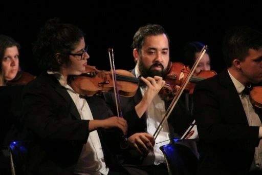 Integrantes da Orquestra Comunitária de São José dos Campos, com vestimentas pretas, tocando um instrumento e em frente a uma parede preta.