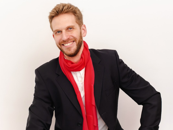 humorista francês paul cabannes posando para foto sorrindo com camiseta branca, blazer preto e cachecol vermelho