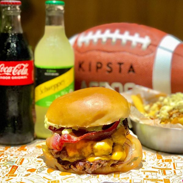 Lanche do PlayOff Burger, uma das opções de hamburger artesanal em SJC (São José dos Campos), no bairro Aquarius; foto mostra um lanche em frente a bebidas, uma porção de batata e uma bola de futebol americano ao fundo.