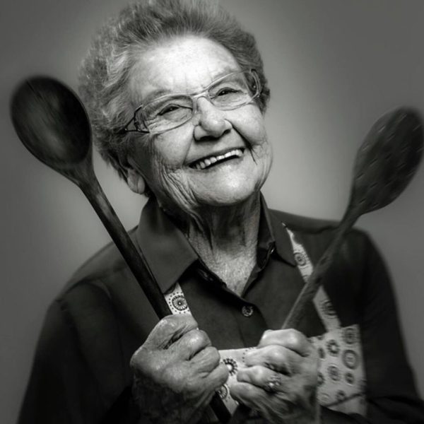 Retrato da Vovó Palmirinha em preto e branco, segurando duas colheres de pau, enquanto sorri para a câmera.