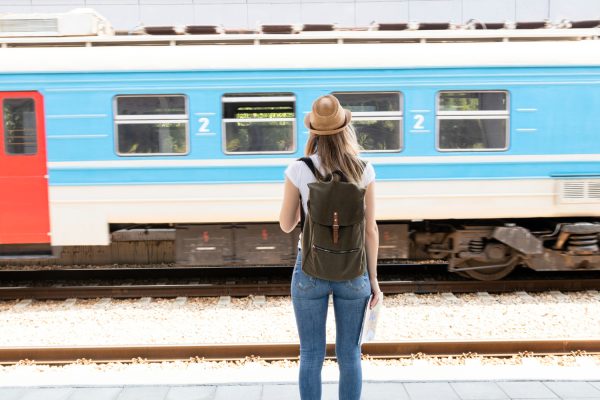 Foto com um trem de linha férrea, ilustrando a matéria sobre a coluna ESG na Prática; foto mostra uma mulher observando um veículo azul com janelas de vidro, enquanto usa um chapéu marrom, camiseta branca e calça jeans.