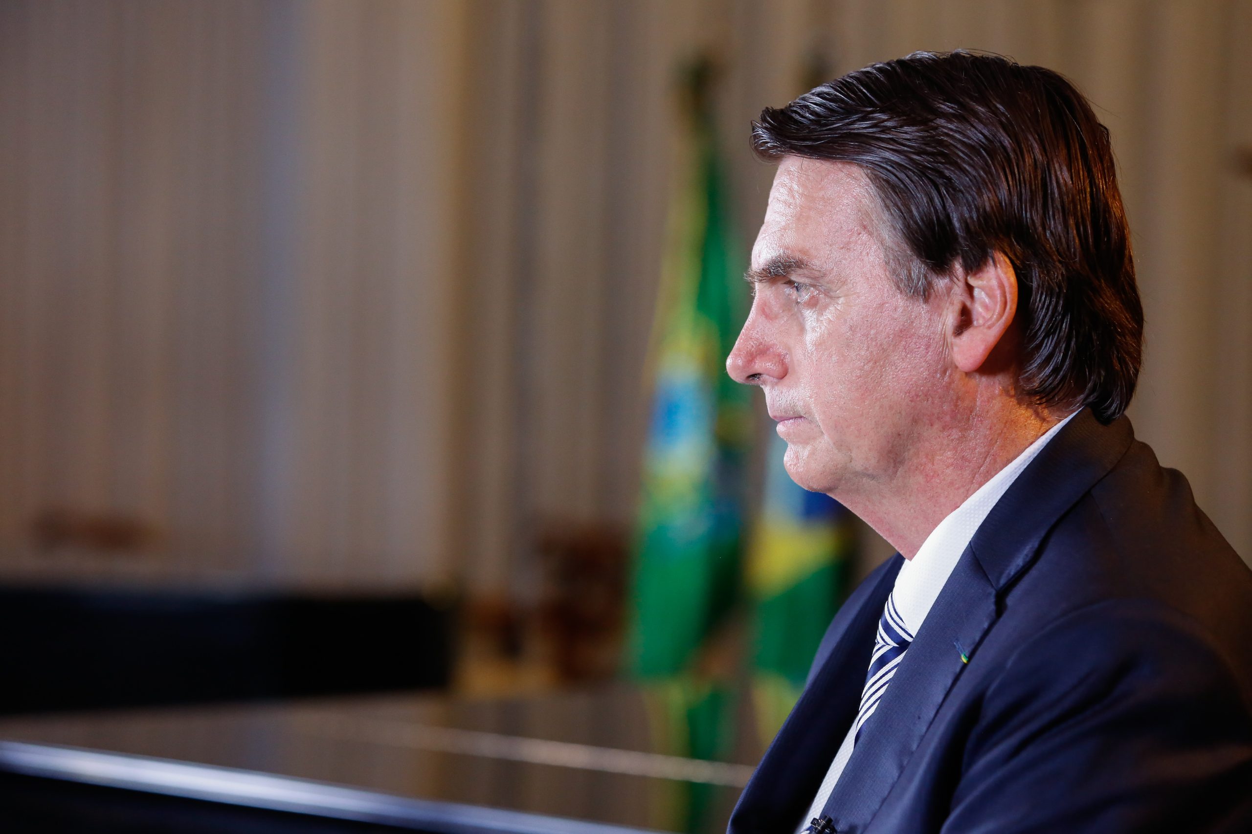 Foto mostra bolsonaro visto de lado, com bandeira do Brasil ao fundo. Julgamento de Bolsonaro será retomado nesta terça (27)
