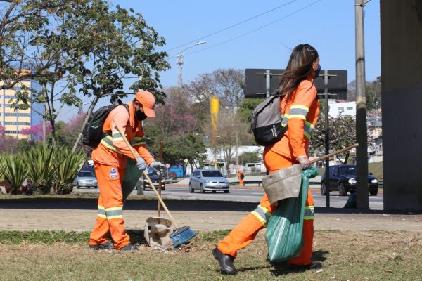 agentes de limpeza em são josé dos campos em serviço nas ruas da cidade. mais à direita, homem com uniforma laranja e boné aparece varrendo e catando lixo com pá, enquanto mais à esquerda, uma mulher, também de uniforme, carrega sacolas de lixo.