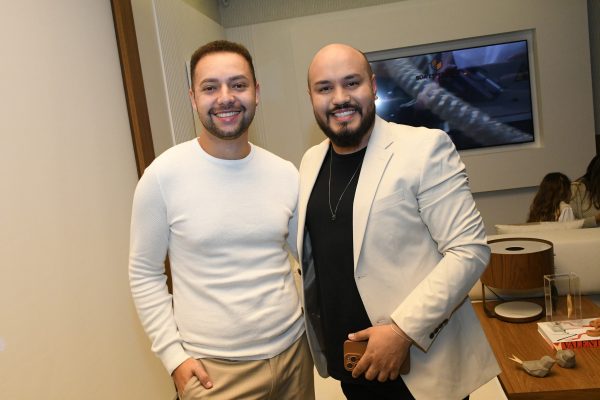 Homem de blusa branca sorrindo ao lado de homem de terno cinza claro.