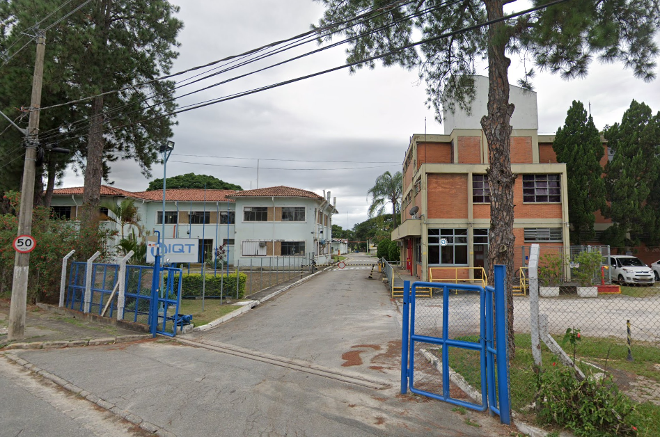 Fachada da indústria de produtos químicos IQT, na qual houve vazamento de um gás. Na fachada, um portão azul, atrás tem um prédio de tijolos laranjas na direita e prédios verdes claros a esquerda.