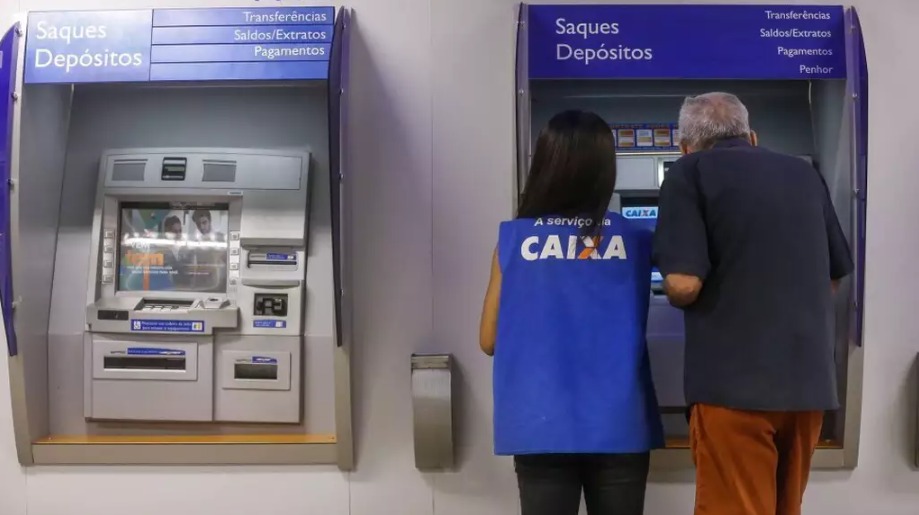 Jovem, ajudando cliente em caixa eletrônico de agência da Caixa Econômica Federal