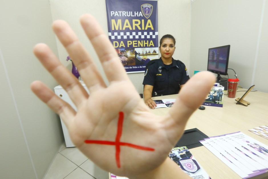 Ao centro, uma mão com um X vermelho e ao fundo, uma policial com a farda da GCM de São José sentada com um cartaz da Patrulha Maria da Penha atrás.