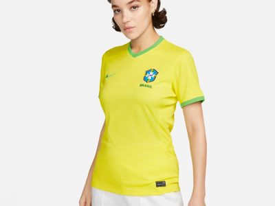 modelo com uniforme 1 da seleção brasileira feminina para a copa do mundo. a camisa é amarela e apresenta uma marca d'água de folhagem tropical por toda a sua extensão, gola em "v" verde e uma barra no final das mangas também na cor verde.
