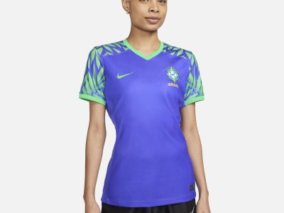 modelo com uniforme 2 da seleção brasileira feminina para a copa do mundo. a camisa é azul e apresenta gola em "v" verde e uma estampa de olhas tropicais das mangas até os ombros também na cor verde.