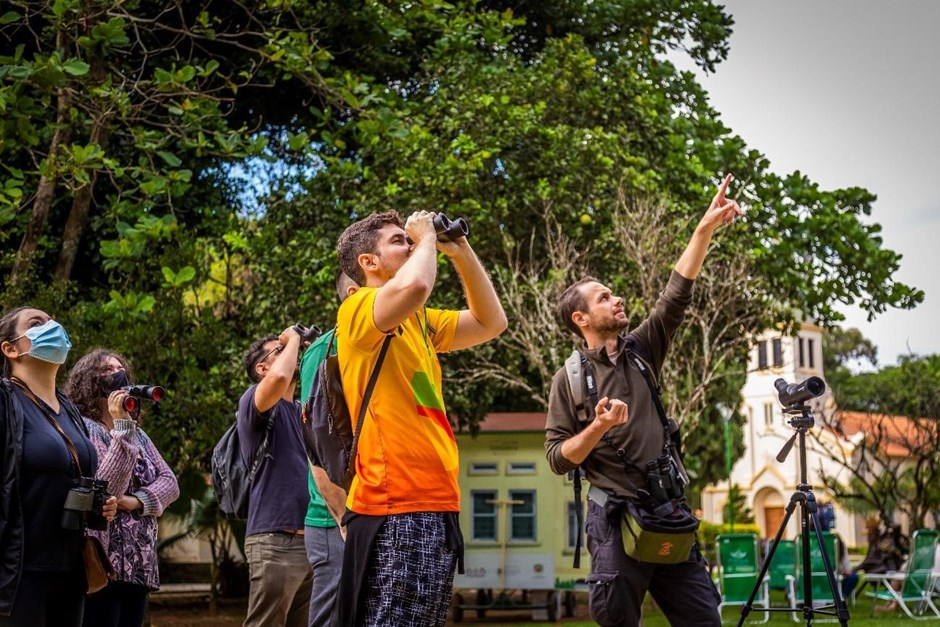 Grupo de pessoas com um homem ao centro observando o céu com um binóculo e outro homem do lado direito apontando para cima