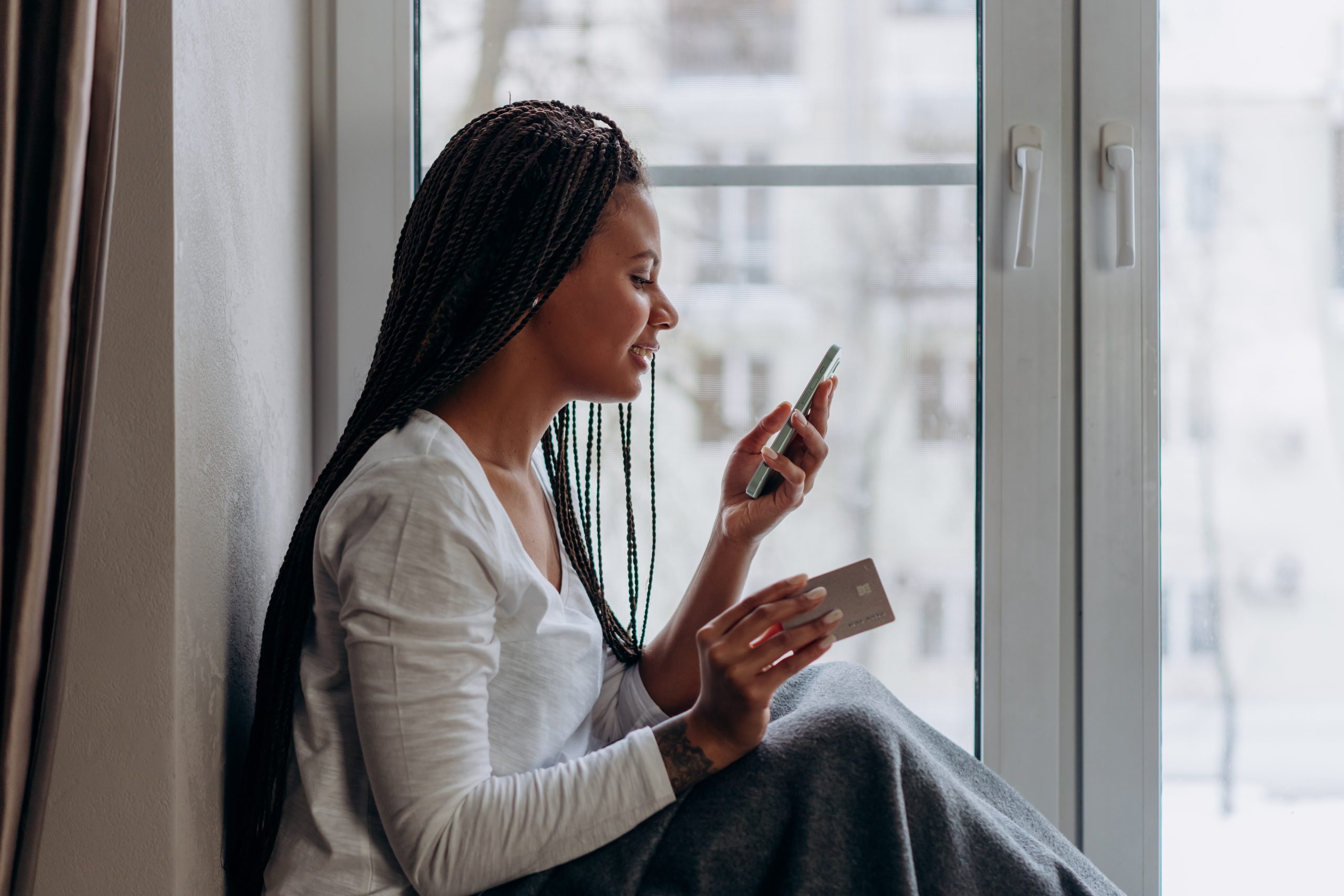 Moça sentada do lado de dentro da janela de seu apartamento, segurando um celular e um cartão de crédito. As novas regras para compras internacionais na internet foram publicadas pelo Ministério da Fazenda