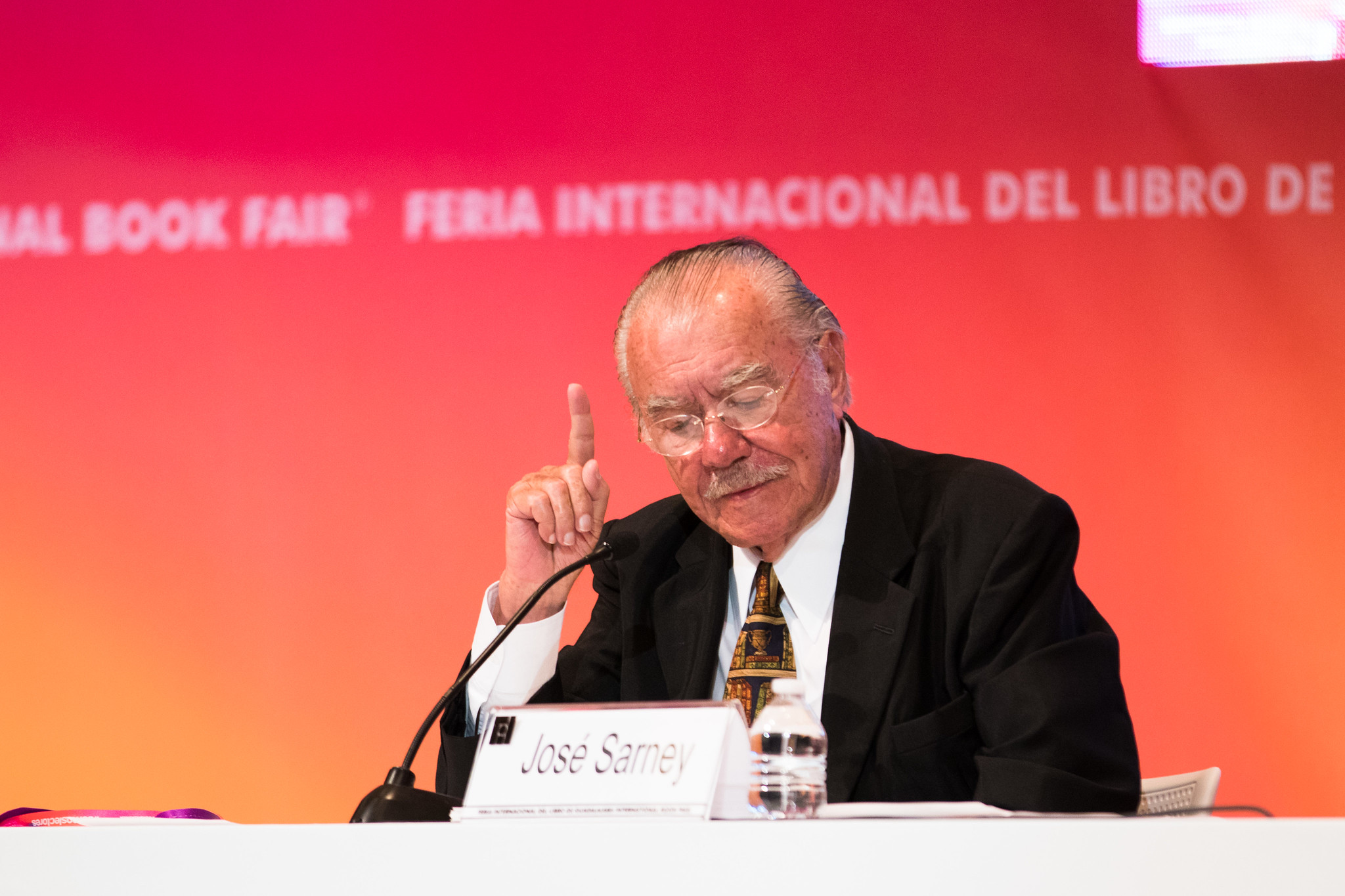 ex-presidente josé sarney, internado no último domingo, em evento internacional de literatura. ele está sentado em banca e aparece com o dedo indicador para cima em posição de contestação.