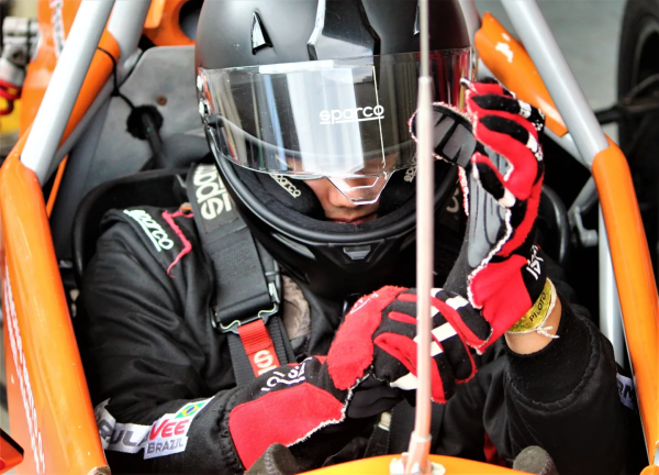 vinicius visto por cima do cockpit. ele veste macacão e capacete pretos e está sentado dentro de carro laranja enquanto arruma luvas e se prepara para corrida