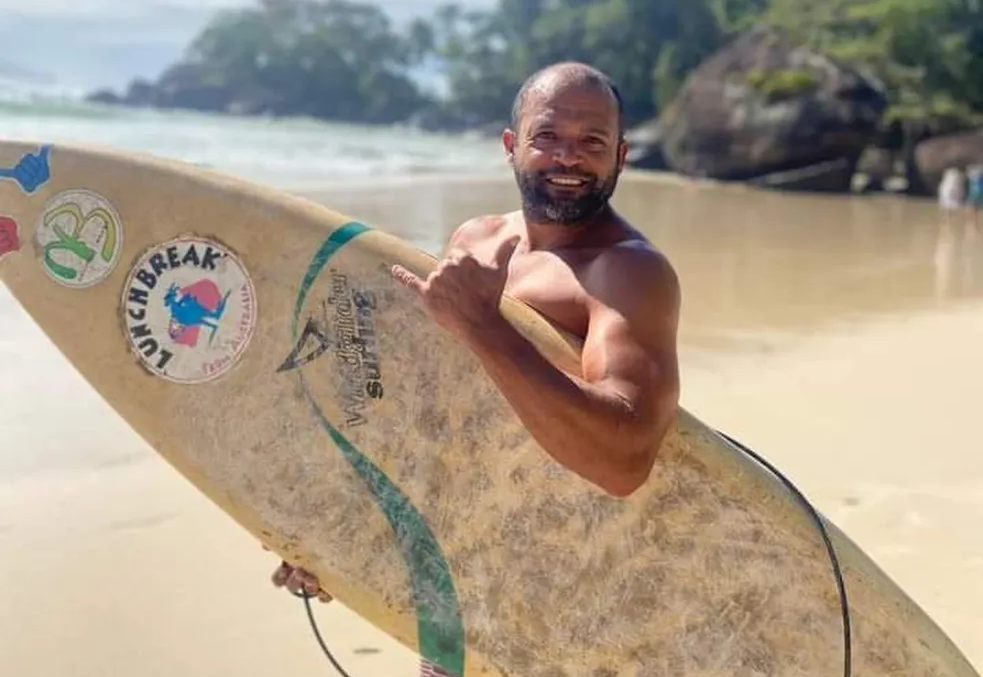 pescador desaparecido em ubatuba, almendes; ele sorri e segura prancha de surfe agarrada ao tronco
