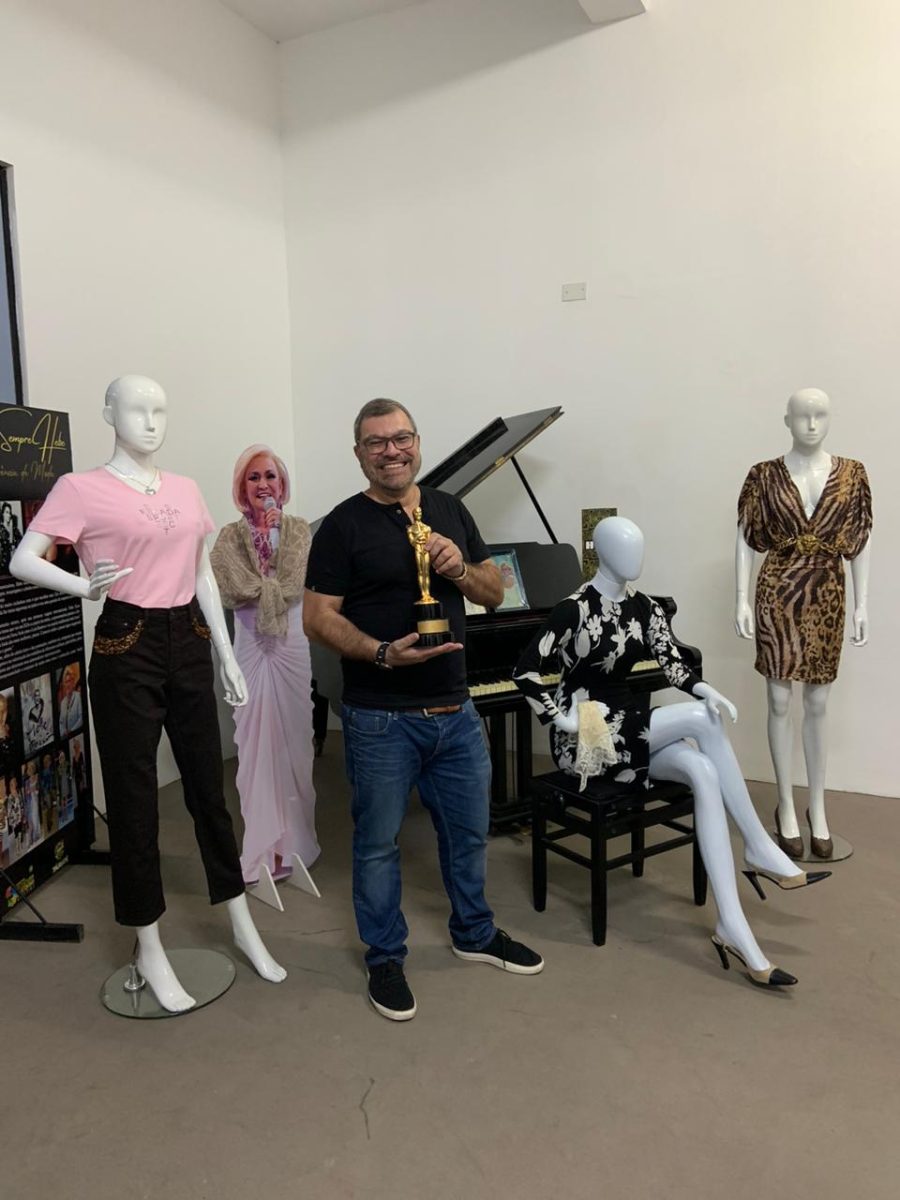 Filho de Hebe Camargo durante visita ao Mistau em Taubaté; ele está ao lado de uma foto e de manequins vestidos de roupas da artista de Taubaté