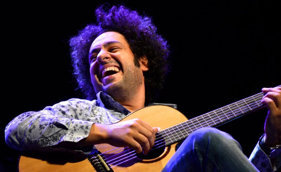 diego figueiredo sentado no palco sorrindo enquanto segura violão no colo