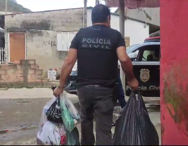 Policial civil carregando sacolas com objetivos apreendidos durante operação que investiga morte de motorista de aplicativo em Ubatuba