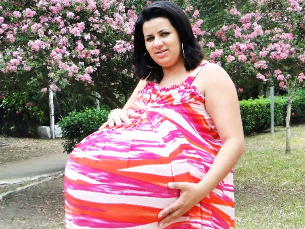 grávida de taubaté em clássica foto com as mãos sobre a barriga em parque com árvores e flores
