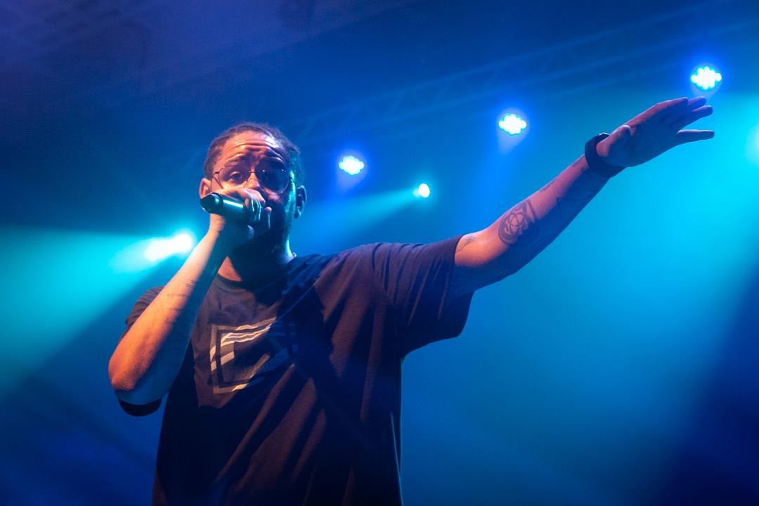 emicida cantando em palco iluminado por hlofotes em azul no sesc de são josé dos campos em 2017; ele abre o braço esquerdo enquanto, com o direito, leva o microfone á boca