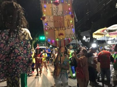 bloco em cuba lá pelas ruas de são josé; mulher à frente carrega estandarte enfeitado