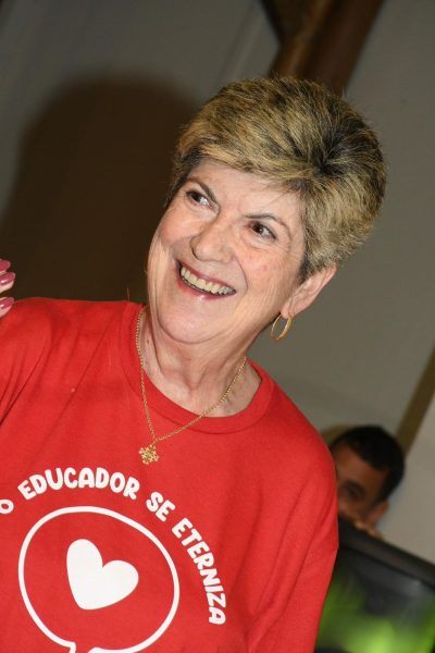 marina ricci, fundadora da escola monteiro lobato, sorrindo; ela é uma mulher branca, de cabelos loiros, curtos e lisos, com franja penteada para o lado e veste camiseta vermelha