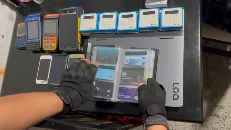 Mãos de Policial Federal (PF) de São José dos Campos, em operação de combate a fraudes bancárias; na imagem, máquinas de cartão estão expostas em uma mesa, o lado de aparelhos celulares