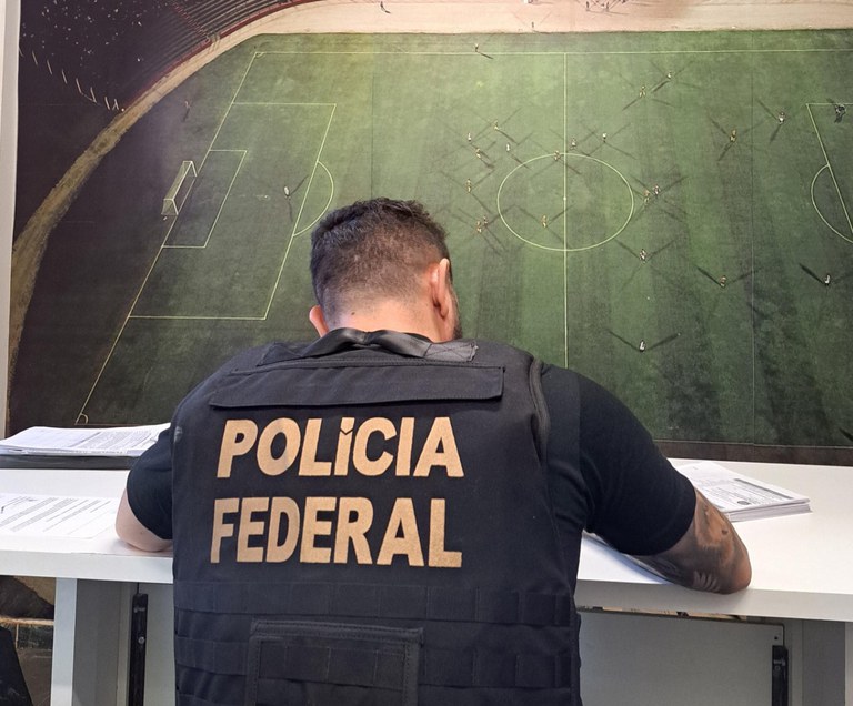 agente da polícia federal de colete anotando coisas em frente à plano de fundo de campo de futebol