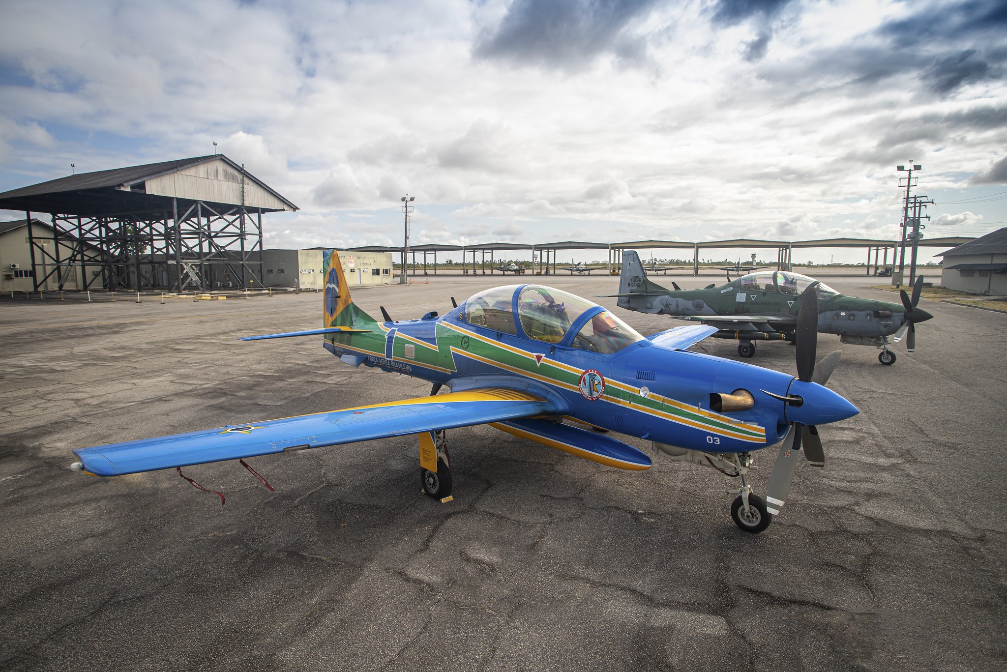 a-29, aeronaves da esquadrilha da fumaça, estacionadas em pátio; uma, mais à frente, tem pintura coma s cores do brasil. a outra, atrás, é camuflada em tons de cinza