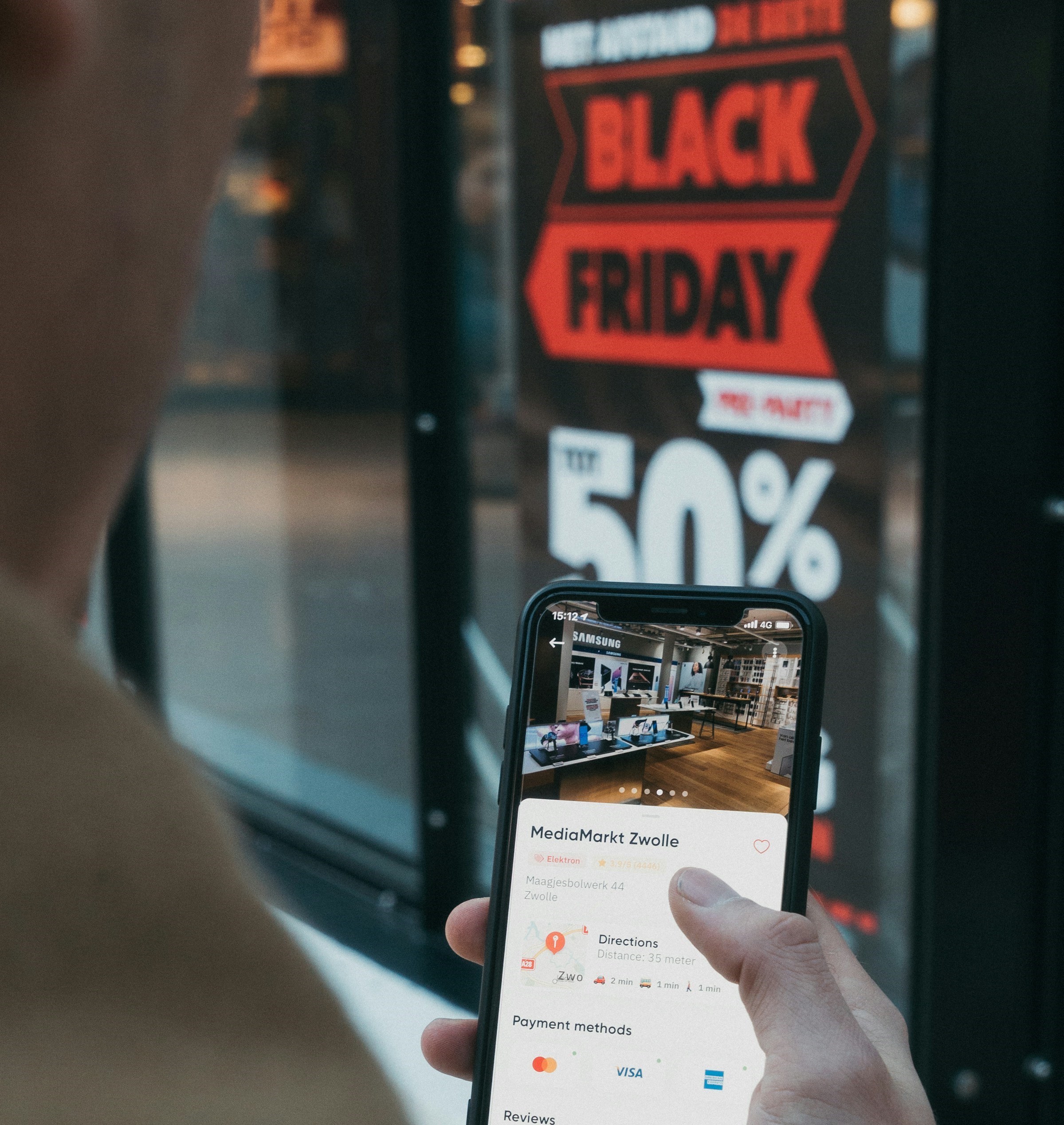 close na tela de um celular realizando compra online; aos fundos, aparece uma vitrine com "black friday 50%"