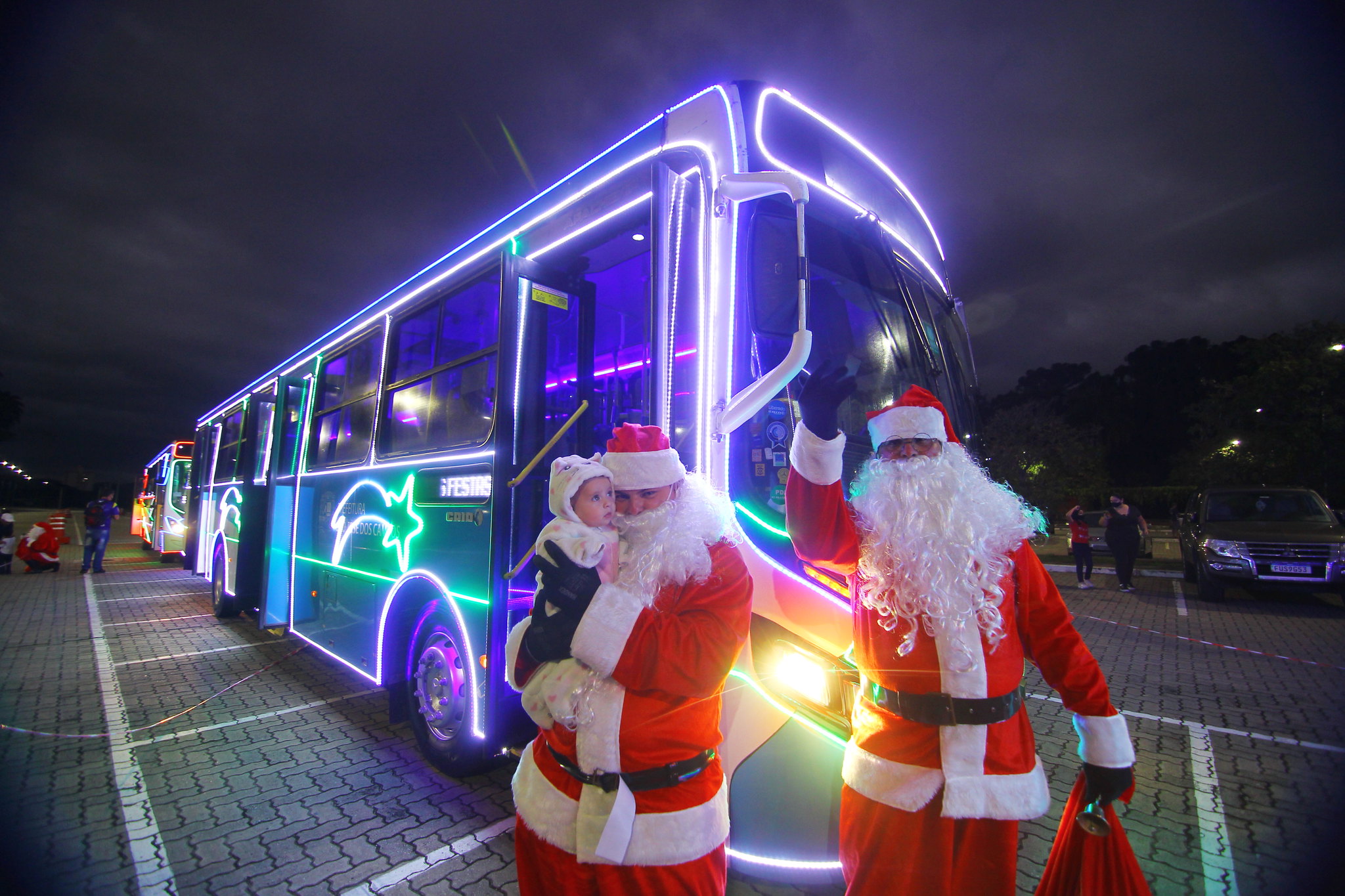 papais noéis, um deles segurando uma criança no colo, em frenta a ônibus da caravana iluminada