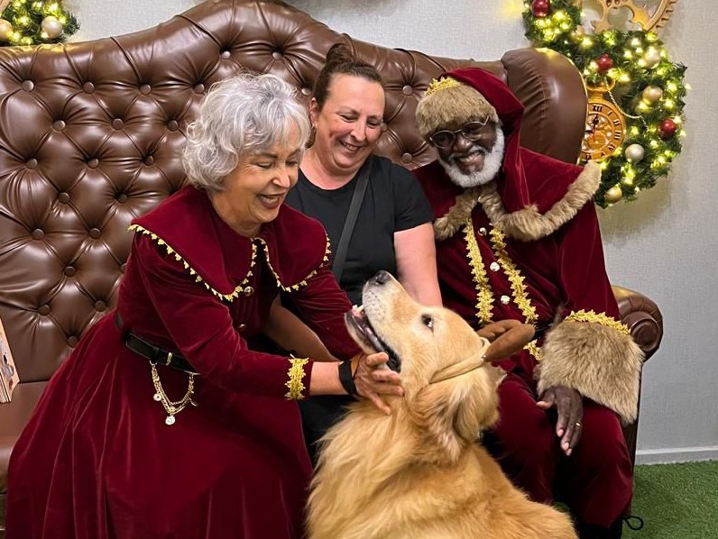 pmamãe noel fazendo carinho no rosto de golden retriever enquanto tutora sorri ao lado de papai noel