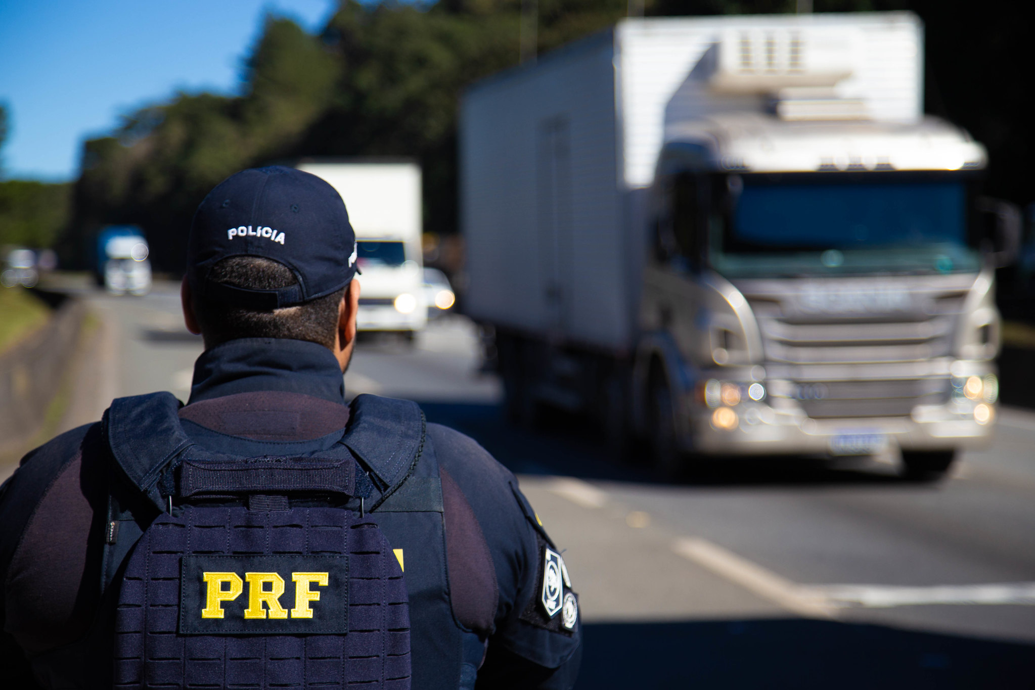 policial da prf na margem de rodovia enquanto passa caminhão