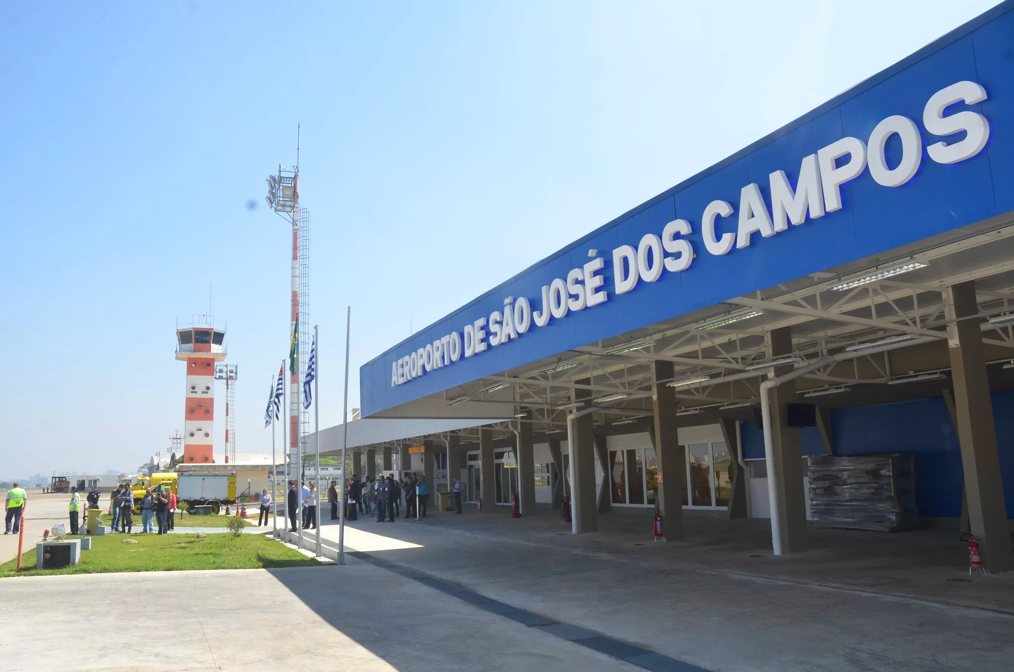 Fachada do Aeroporto de SJC, com céu azul, placa, área verde e hall na frente.