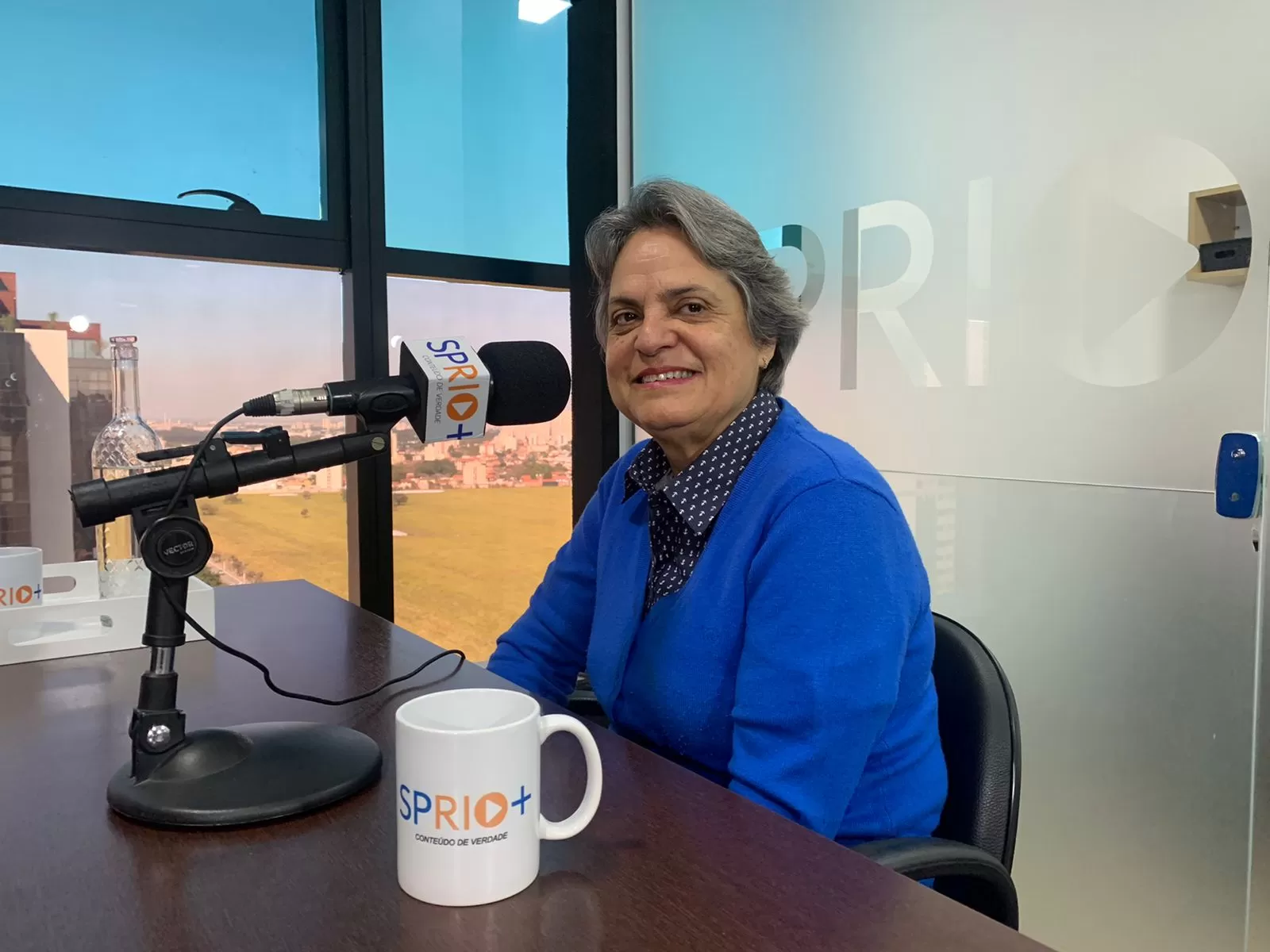 Retrato da Flávia Camargo sentada na mesa da SP RIO+, com uma caneca, um microfone e São José dos Campos ao fundo.