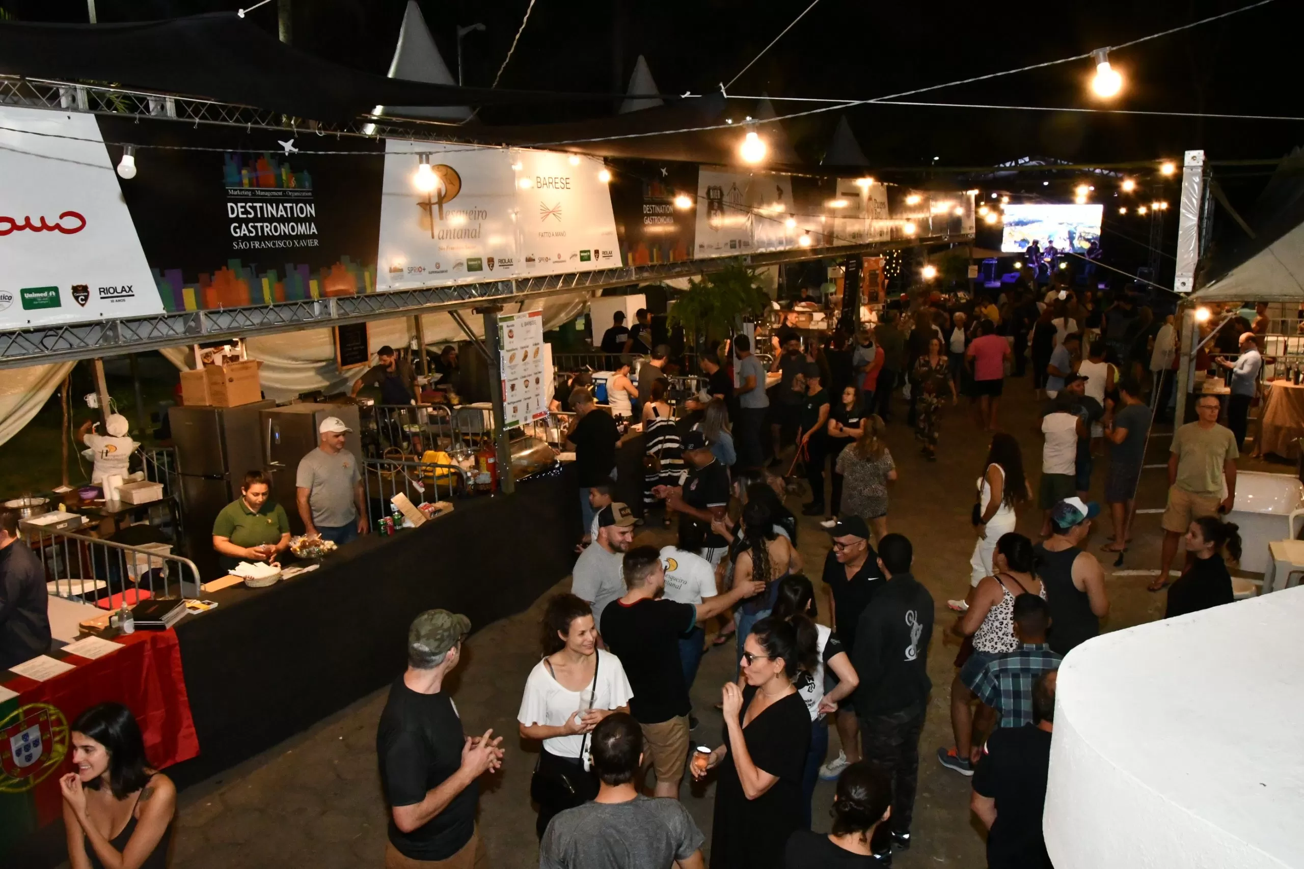 Pessoas conversando, com stands em volta, luzes em cima, comidas expostas, durante o Festival Destination Gastronomia em São Francisco Xavier.