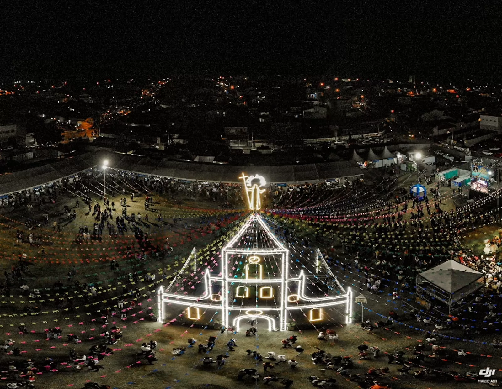 Vista aérea de uma Festa de João, com uma igreja iluminada, com bandeirinhas juninas coloridas em volta, diversas pessoas em volta e a cidade ao fundo. O local é um dos pontos turísticos de Caçapava.