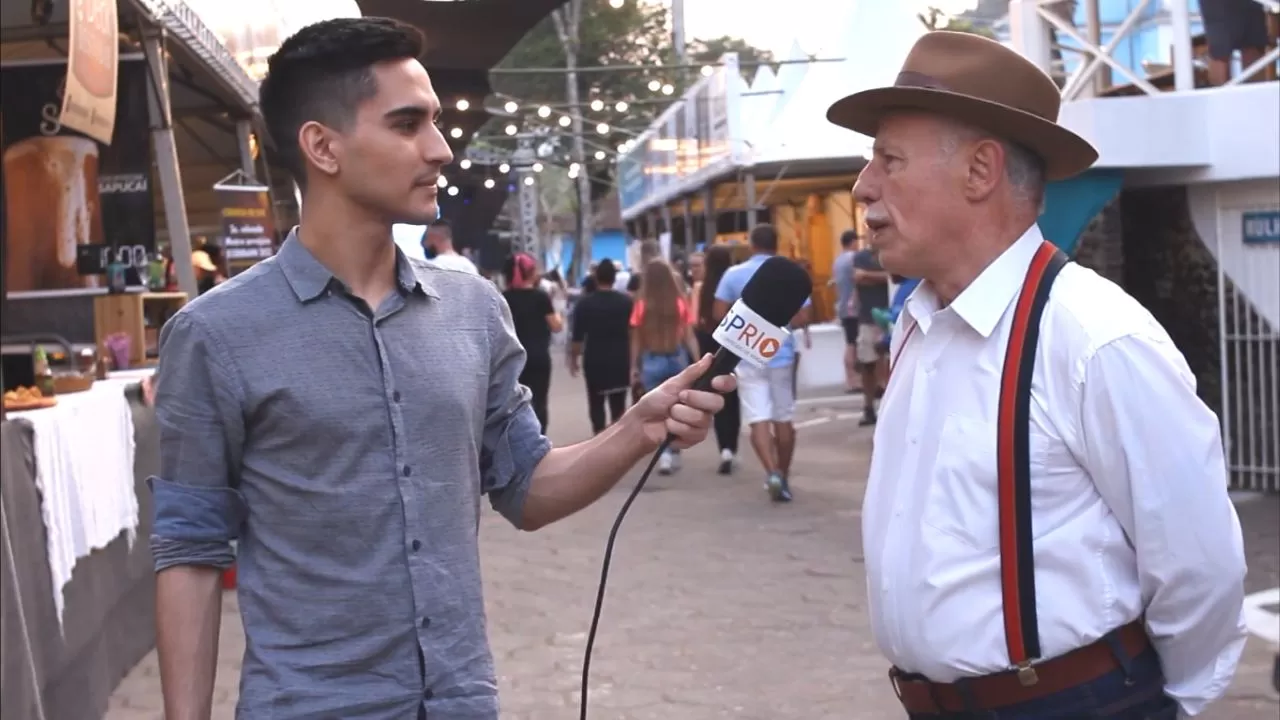 Jornalista Matheus Andrade segurando um mirofone, apontando para o entrevistado Carlito, à direita, que veste uma camisa branca, um suspensório vermelho e um chapéu, com o evento ao fundo.