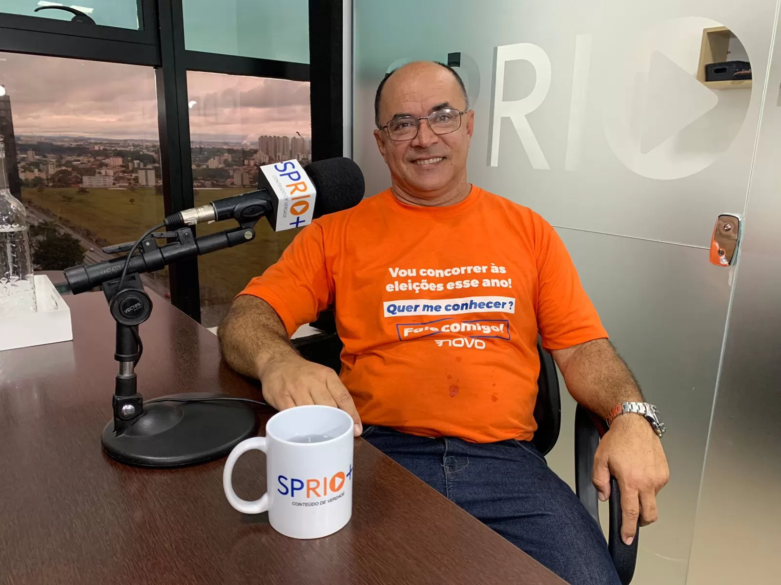 Ju Guimarães, de Jacareí, candidato do Partido Novo a deputado estadual, sentado na cadeira da SP RIO+, com um microfone na mesa e a cidade ao fundo.