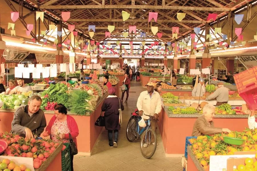 Mercado Municipal de Caçapava, com bandeirinhas no teto, pessoas circulando, frutas expostas em balcões, além de verduras.