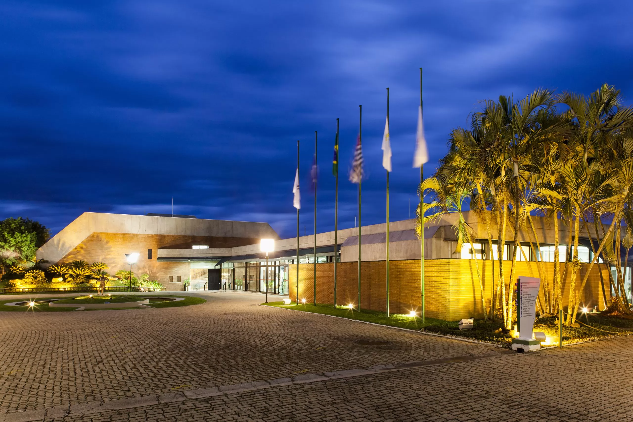 Fachada do Sesc Taubaté, com luzes, árvores e bandeiras em volta; com um céu azul em cima.