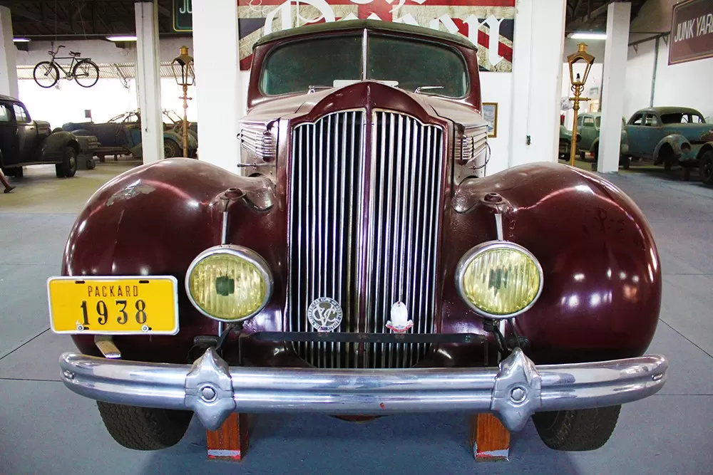 Carro bordô, modelo Packard 1938, exposto no Museu Roberto Lee, de frente para a câmera, com uma placa amarela e o museu ao fundo, com menos detaque.