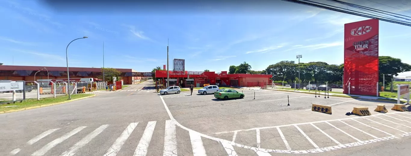 Entrada da fábrica Nestlé, em Caçapava, com destaque para a larga rua e o prédio em vermelho, com placas grandes com o nome, além de carros estacionados em frente.