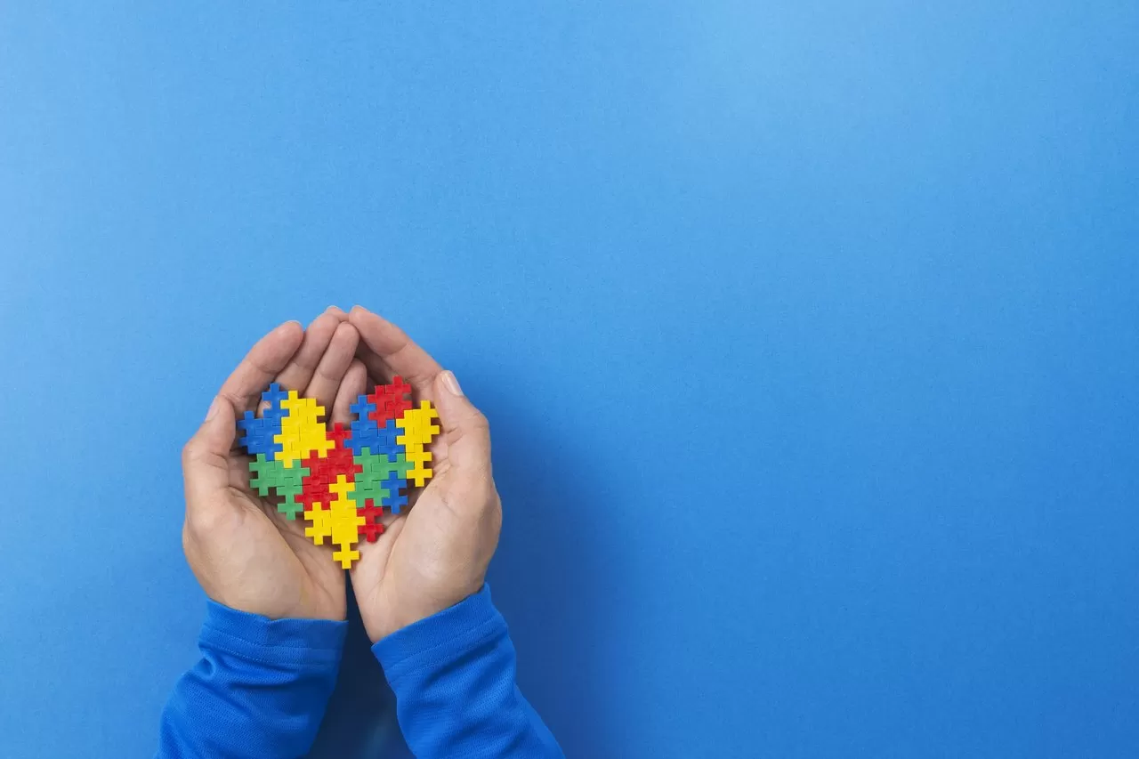 Mão segurando um coração de quebra cabeças coloridos, símbolo do autismo, com um fundo azul.